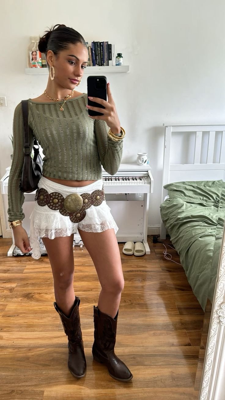 Jovem mulher vestindo uma blusa verde de manga comprida, saia curta branca de renda com cinto largo decorado, e botas estilo cowboy marrons. Acessórios incluem brincos grandes, colar e pulseiras douradas.