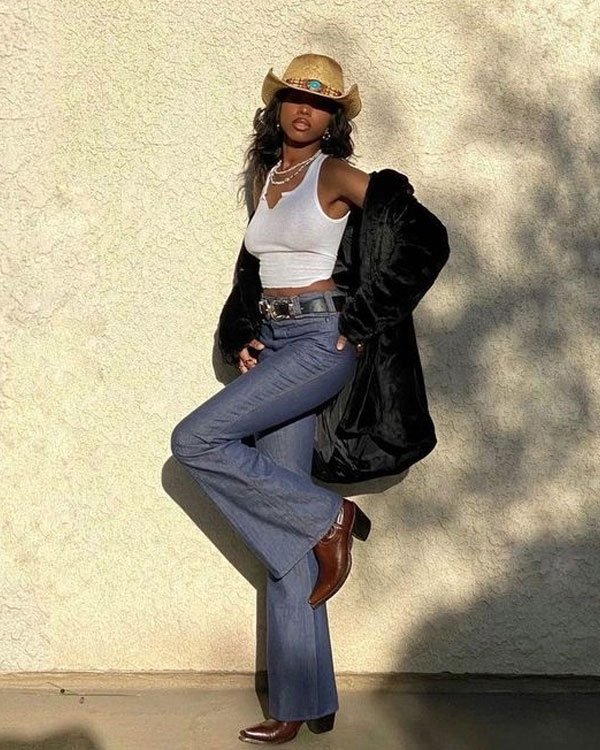 Pessoa usando chapéu de palha com detalhes em pedras, top branco cropped, calça jeans boca de sino, cinto com fivela grande e casaco preto. Completa o look com botas de salto baixo e colares acessórios.