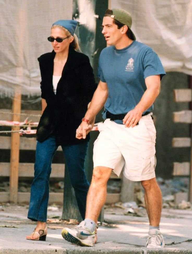 Casal andando de mãos dadas. Ela usa óculos de sol, camiseta preta, jeans e sandálias de salto. Ele veste boné verde virado para trás, camiseta azul, bermuda branca e tênis esportivo.