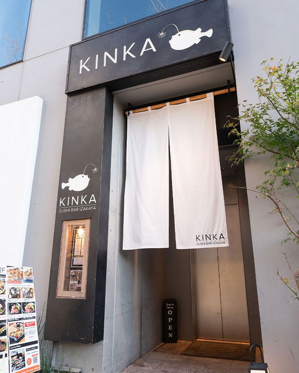 Restaurante Kinka Sushi Bar Izakaya, fachada minimalista e moderna em tons de cinza e branco. Entrada com cortina tradicional japonesa noren, decoração simples e elegante. Atmosfera contemporânea e atraente para os amantes da culinária japonesa.