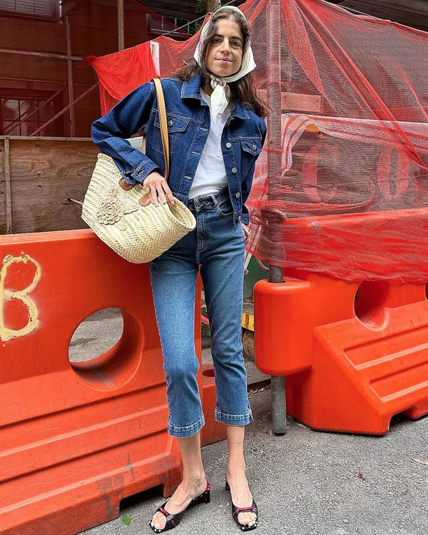 Pessoa vestindo uma jaqueta jeans combinada com calça jeans cropped. Camiseta branca por baixo, lenço na cabeça e bolsa de palha artesanal. Os acessórios incluem sandálias de tiras e uma máscara protetora no rosto. Look casual e tendência retrô.