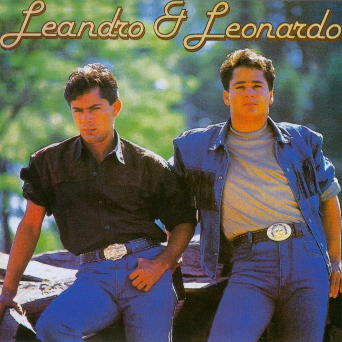 Imagem de dois homens vestindo jeans, cinto com fivela grande e camisa de botão com manga dobrada. Um usa camisa preta e segura algo, o outro usa camisa branca com uma jaqueta jeans com franja. Estilo casual inspirado na moda country dos anos 90.