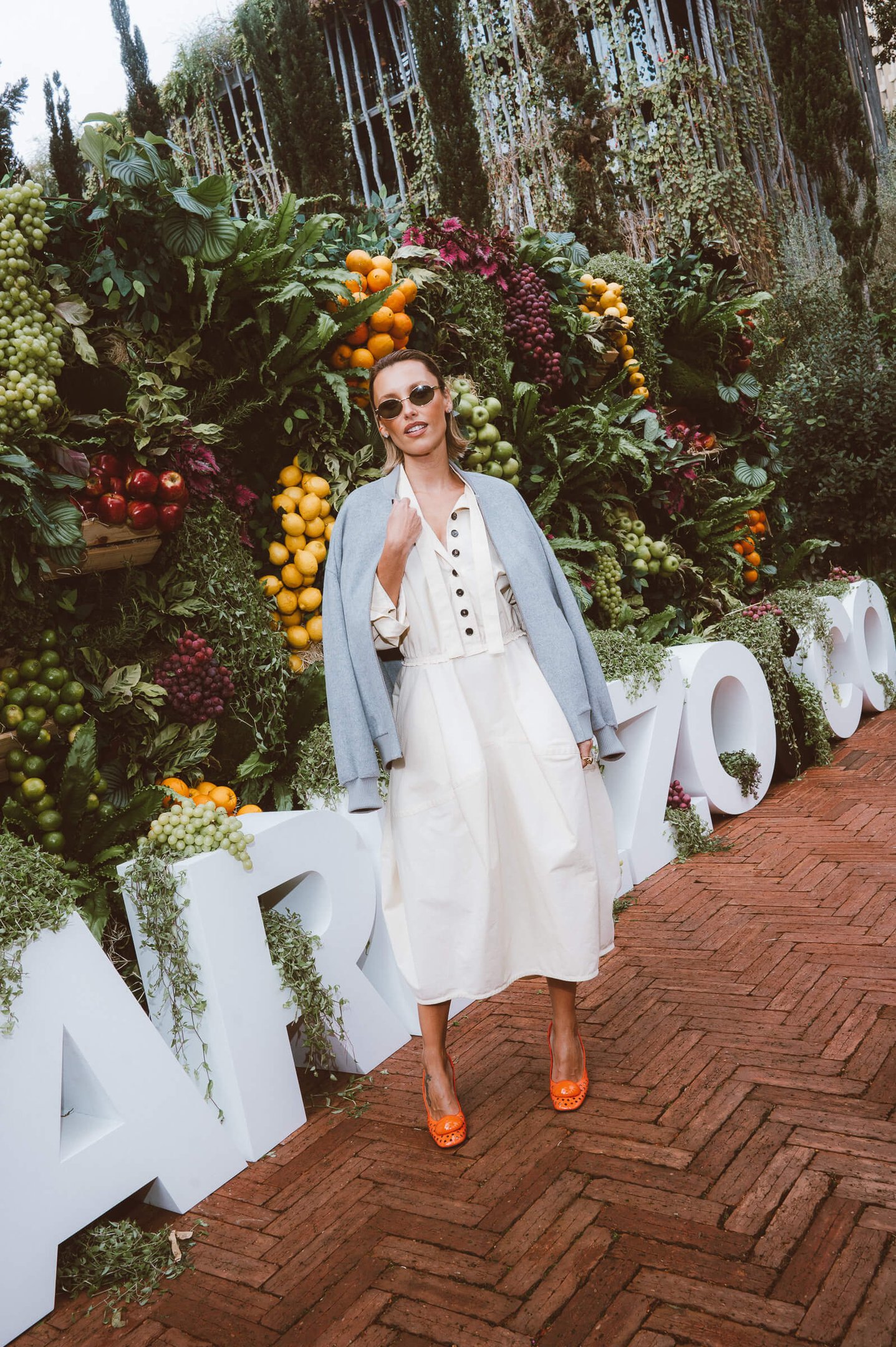 Mulher vestindo um vestido branco de estilo midi com abotoamento frontal, sobreposto com um casaco leve azul claro. Ela complementa o look com sapatos laranja e óculos de sol redondos. O fundo é uma parede de frutas frescas coloridas.