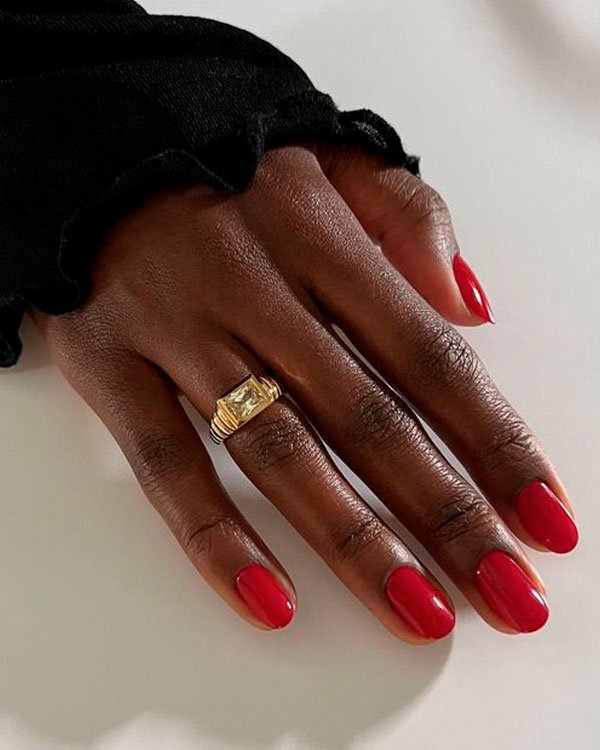 Mão feminina com unhas pintadas de esmalte vermelho, usando um anel dourado com pedra retangular clara. Manga preta de blusa com detalhe em babado.