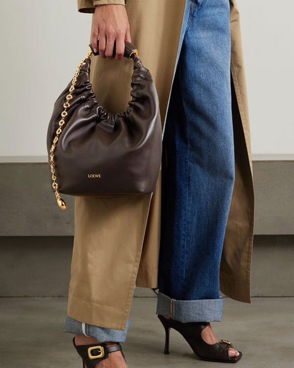 Look moderno com trench coat bege, calça jeans larga e dobrada na barra e sandálias de salto alto. Bolsa de couro marrom com alça de corrente dourada, complementando o visual elegante e casual.