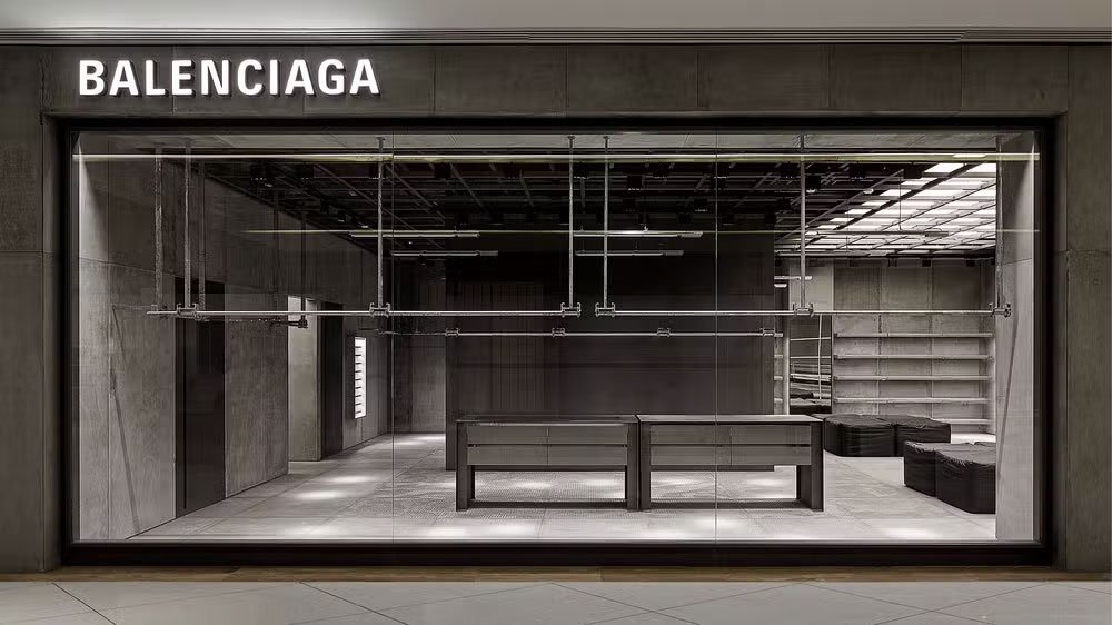 Fachada de loja da Balenciaga com design minimalista e industrial. Espaço interno com tons de cinza e iluminação moderna, refletindo tendências contemporâneas de moda e arquitetura.