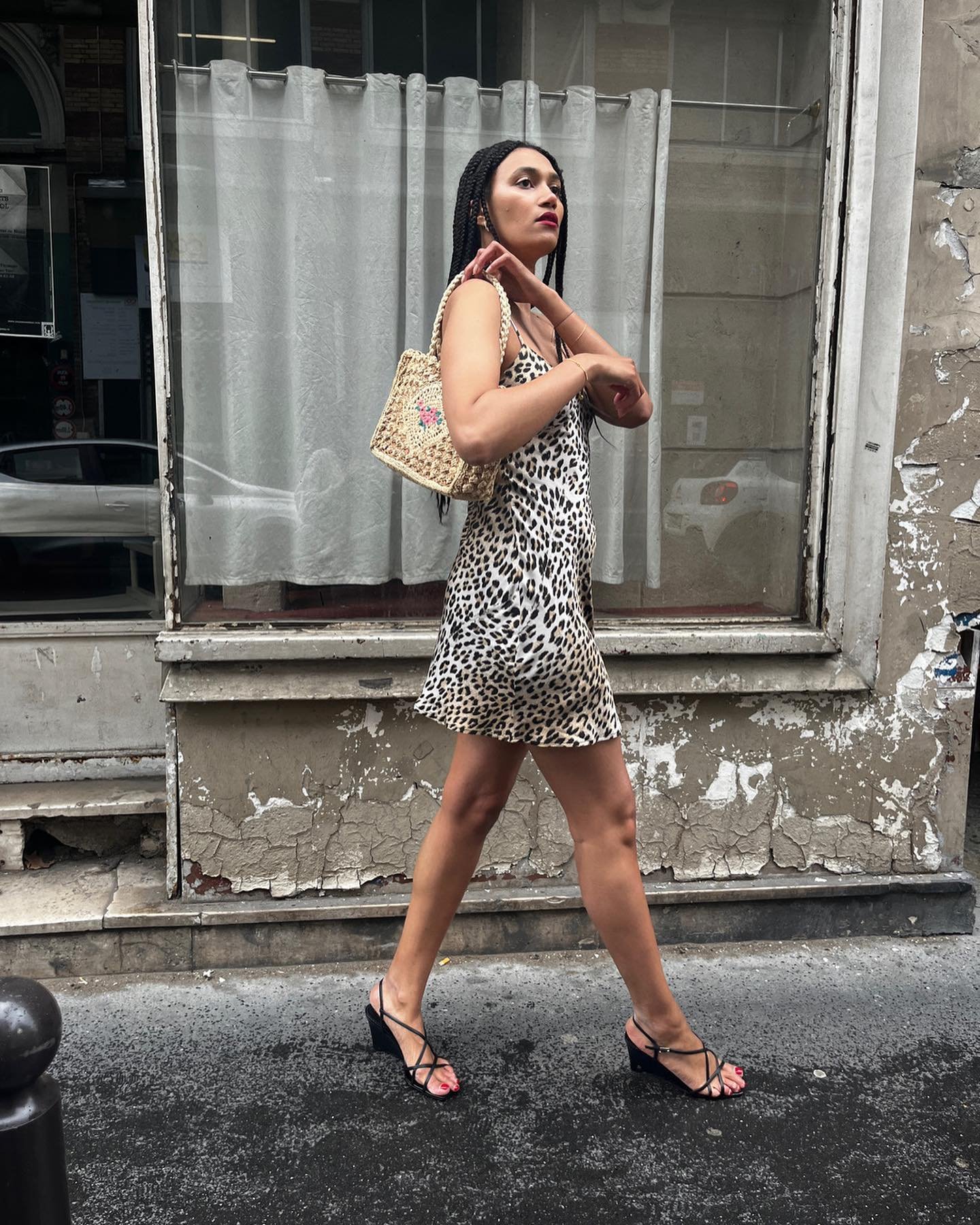 Mulher caminhando pela rua usando um vestido com estampa de oncinha e sandálias pretas de salto. Bolsa de palha pequena e penteado com tranças longas. Look moderno e casual com estilo urbano e influências da moda em animal print.