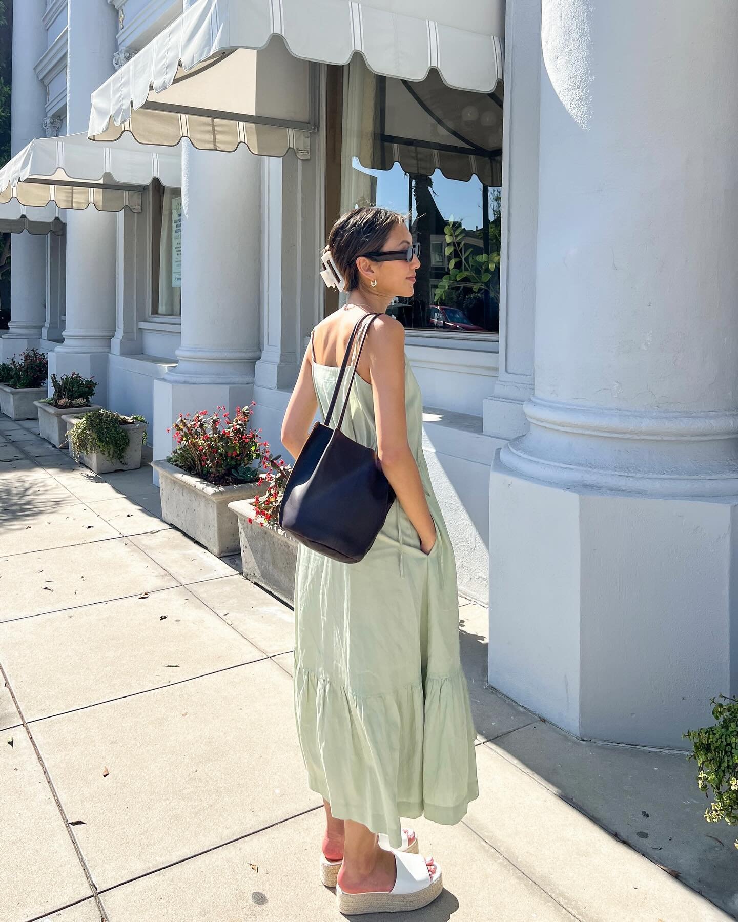 Mulher vestindo vestido midi verde claro com alças finas, sandálias plataforma brancas e bolsa preta de ombro. Acessórios incluem óculos de sol e prendedor de cabelo. Estilo casual e elegante, ideal para o verão.