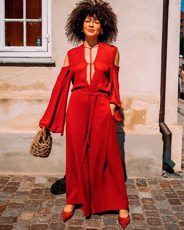 Mulher vestindo um macacão vermelho vibrante com ombros à mostra e mangas longas. Ela segura uma bolsa de palha trançada, adicionando um toque rústico ao look. Completa o visual com sapatos de salto baixo também vermelhos.