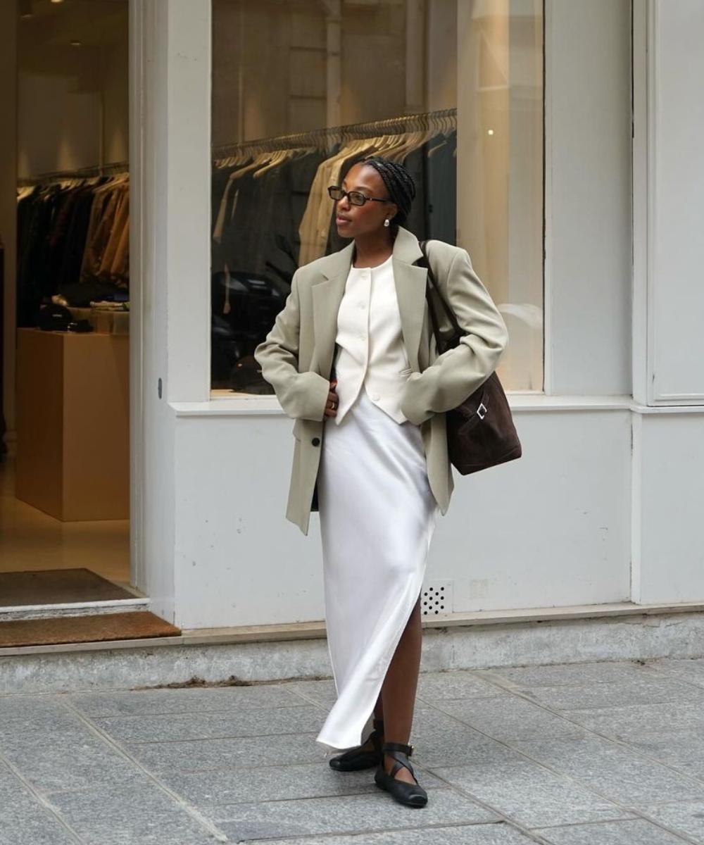 Mulher com estilo clássico e elegante, vestindo blazer oversized bege, vestido branco de cetim, sapatos pretos de tiras e bolsa marrom. Completa o visual com óculos, brincos simples e tranças. Moda atemporal com toques modernos e sofisticados.