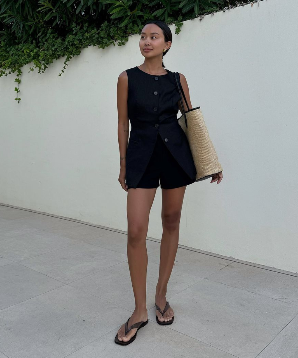 Imagem de uma mulher vestindo um conjunto preto de blusa sem mangas e shorts curtos, com chinelos pretos. Ela carrega uma bolsa de palha grande, complementando um estilo casual e elegante para o verão.