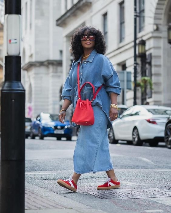 Mulher vestindo look casual e elegante, composto por conjunto jeans oversized. Acessórios incluem bolsa vermelha e tênis combinando, além de óculos de sol grandes. Tendência urbana com estilo confortável e moderno.