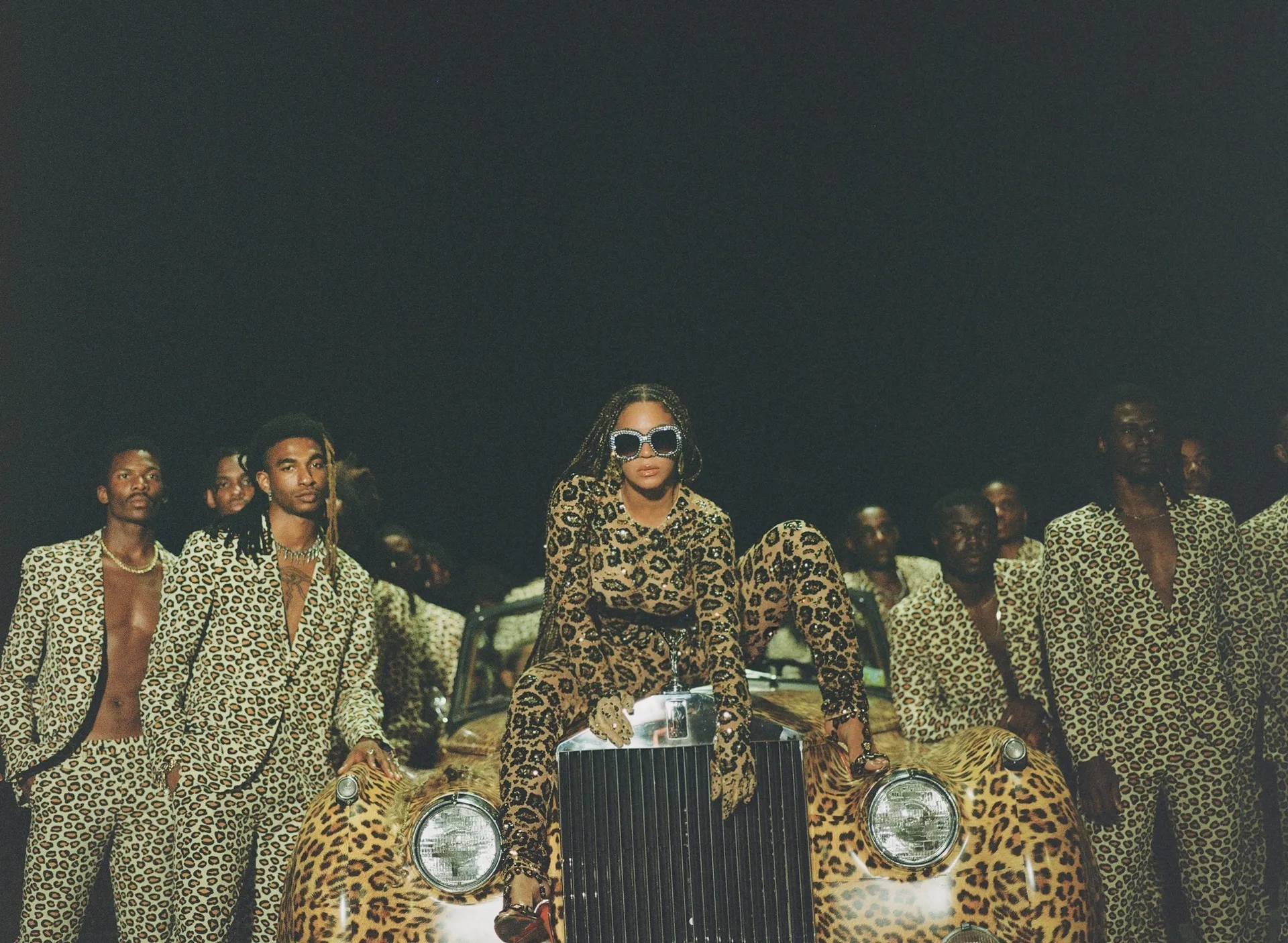 Imagem de pessoas vestindo roupas estampadas de oncinha, destacando uma mulher sentada sobre um carro com a mesma estampa, usando óculos de sol grandes. Looks da Beyoncé.