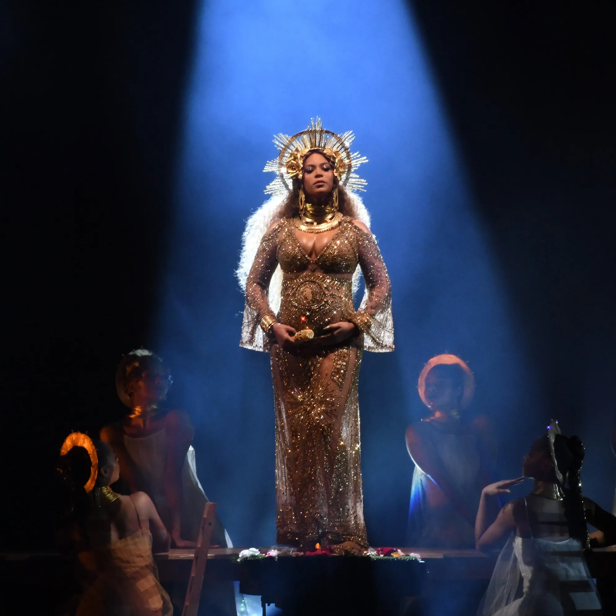 Imagem de uma mulher vestindo um longo vestido dourado com transparências, adornado com joias e uma coroa radiante na cabeça. Looks da Beyoncé.