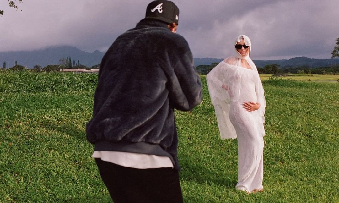 Pessoa ao ar livre usando um vestido branco fluido e fino, com óculos escuros e um véu cobrindo a cabeça. Atrás, pessoa com jaqueta de pelúcia preta, boné e camiseta branca. Looks de grávida Hailey Bieber.