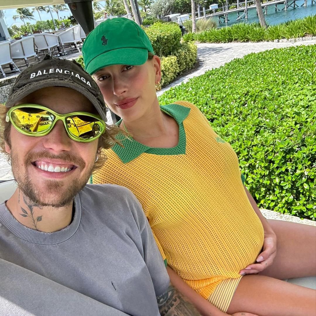 Casal em ambiente externo. Homem usa óculos de sol esportivos amarelos, boné preto Balenciaga e camisa cinza. Mulher veste boné verde, blusa amarela com detalhes verdes e shorts listrados. Estilo casual e despojado. Tendências de moda com marcas e cores vibrantes.