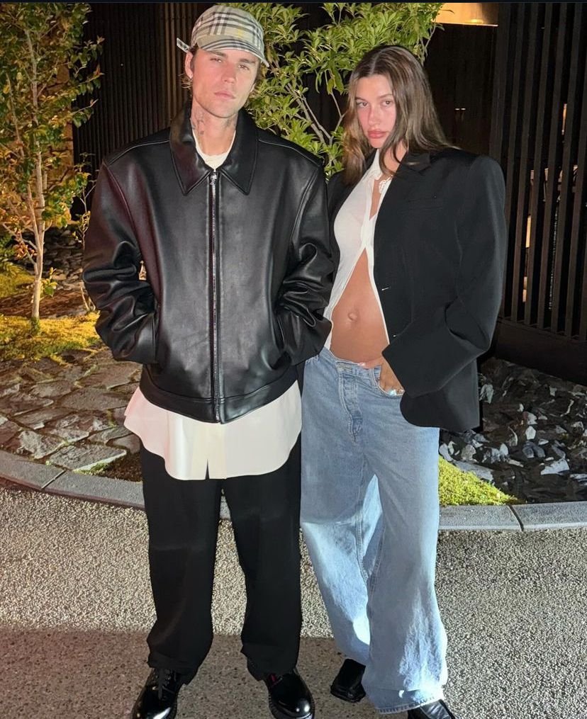 Casal com estilo moderno: Homem veste jaqueta de couro preta, camisa longa branca, calça preta larga e boné xadrez. Mulher usa blazer oversized preto, top branco curto e calça jeans larga. Look despojado e contemporâneo.