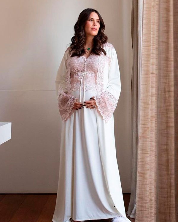 Mulher grávida vestida com robe de cetim branco com detalhes em renda rosa, mangas largas e joias discretas. Look confortável e sofisticado, combina tendências de moda íntima e maternidade.