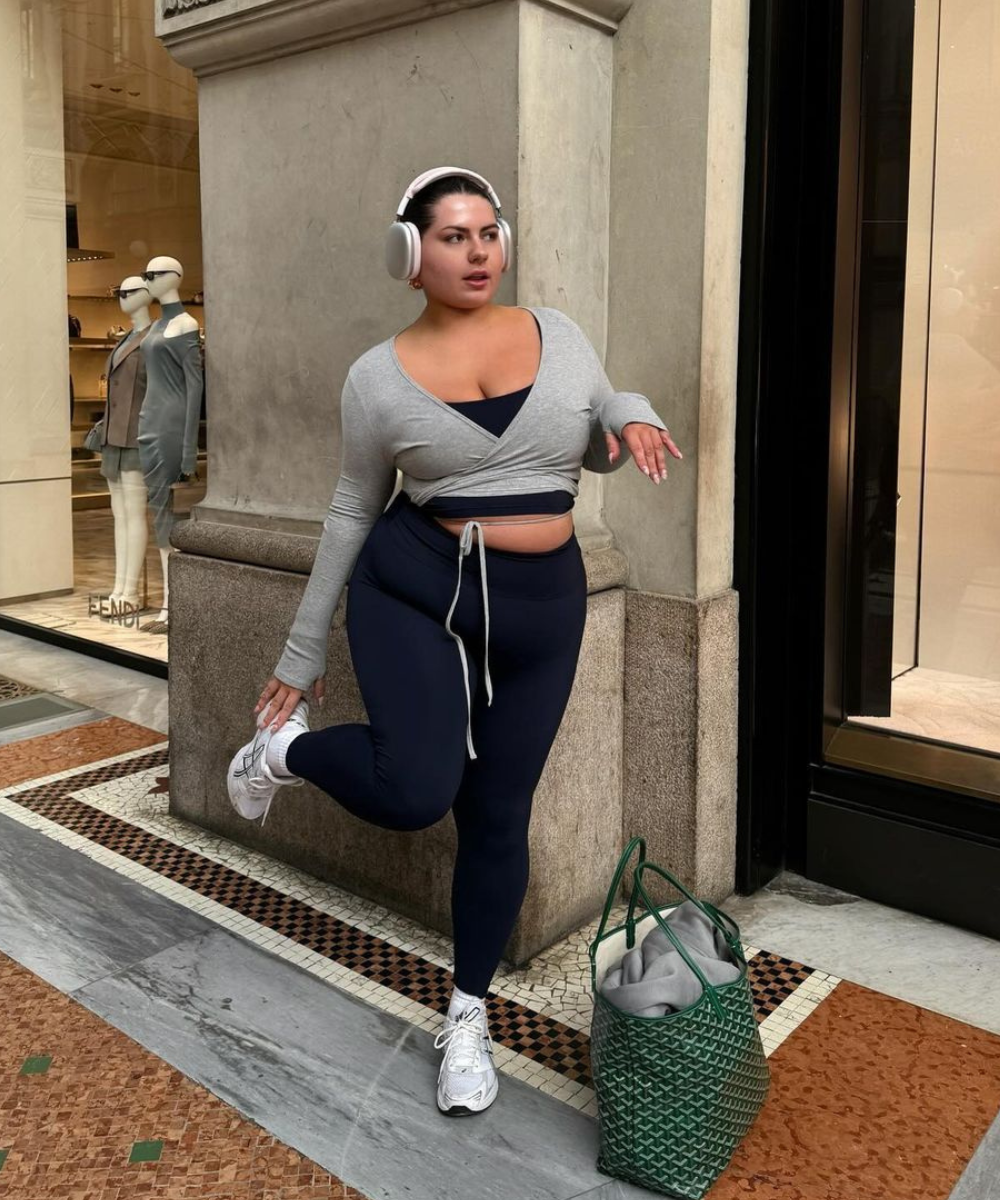 Pessoa com fones, usando cropped cinza de manga longa e legging preta. Look esportivo e casual, combinando com tênis branco e bolsa verde estilo tote. Tendência athleisure, popular para conforto e moda do pilates.