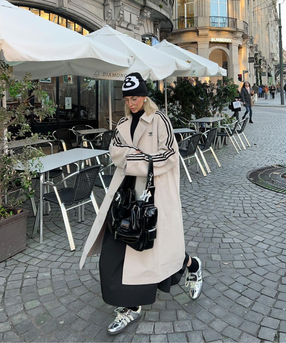 Pessoa usando casaco beige com listras pretas nas mangas, gorro preto com número 8, óculos de grau, calça preta, tênis metalizados e bolsa preta volumosa. Combinação de estilo esportivo e chique urbano, destacando-se por acessórios modernos e ousados.