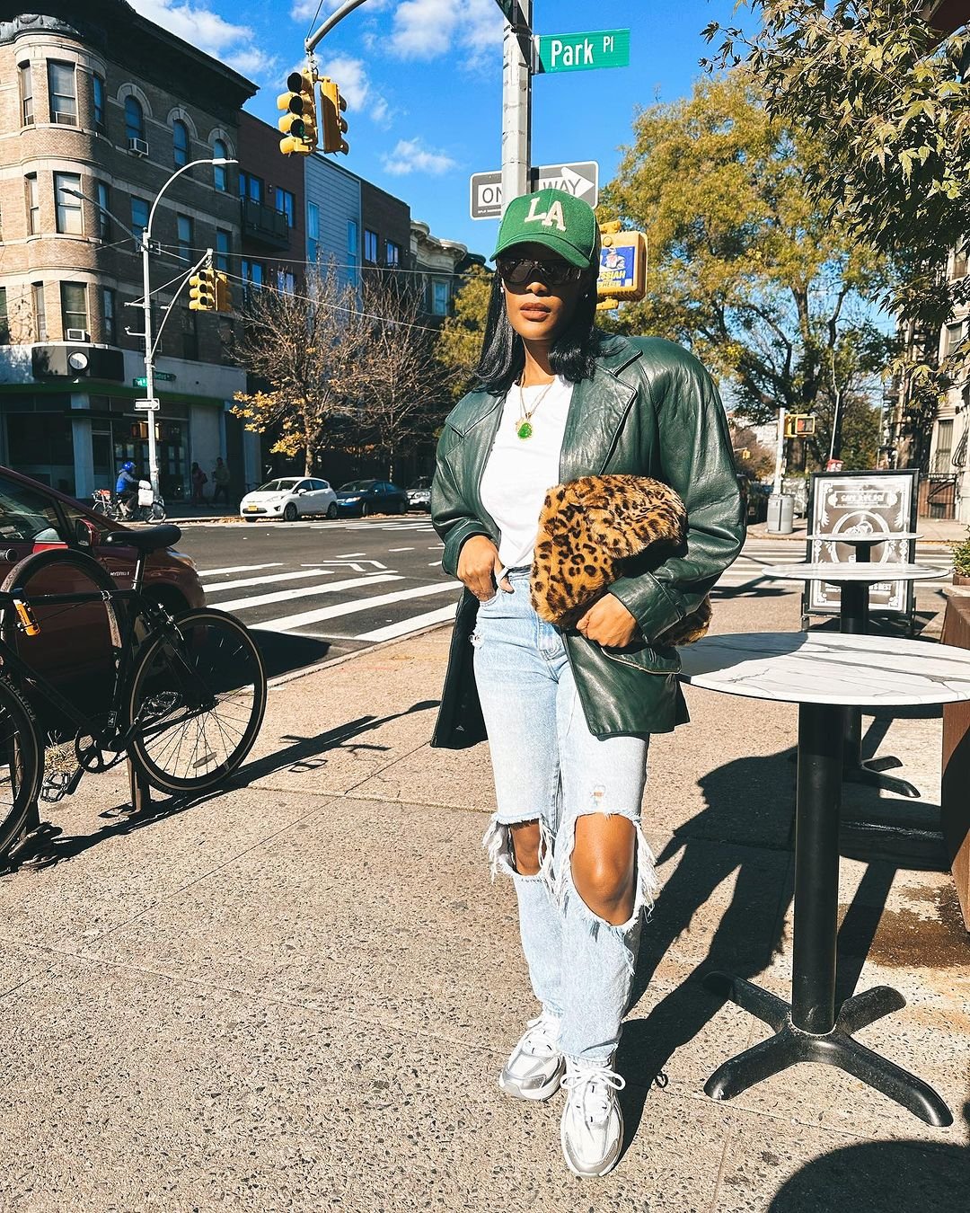 Pessoa com boné verde LA, óculos de sol, jaqueta de couro verde, camiseta branca, jeans rasgados e tênis prata. Carrega bolsa de pelúcia com estampa de leopardo. Estilo casual e despojado.