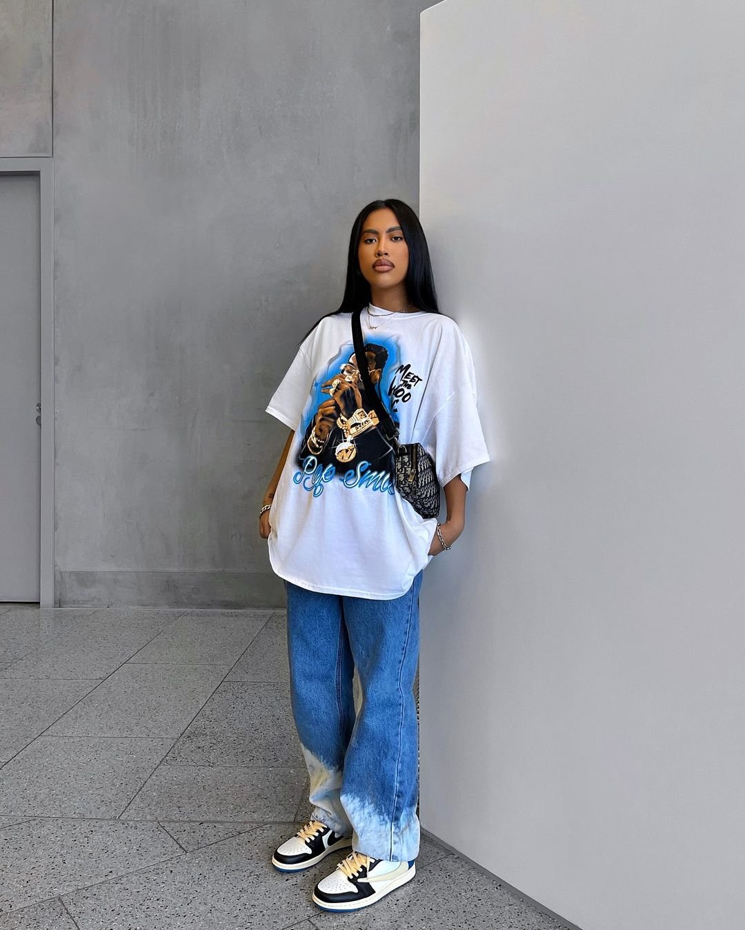 Mulher moderna com camiseta gráfica oversized, calça jeans mom fit de lavagem clara, tênis de cano médio estilo retrô e bolsa transversal preta. Look casual e descolado, perfeito para ocasiões urbanas e destacando tendências de moda streetwear.