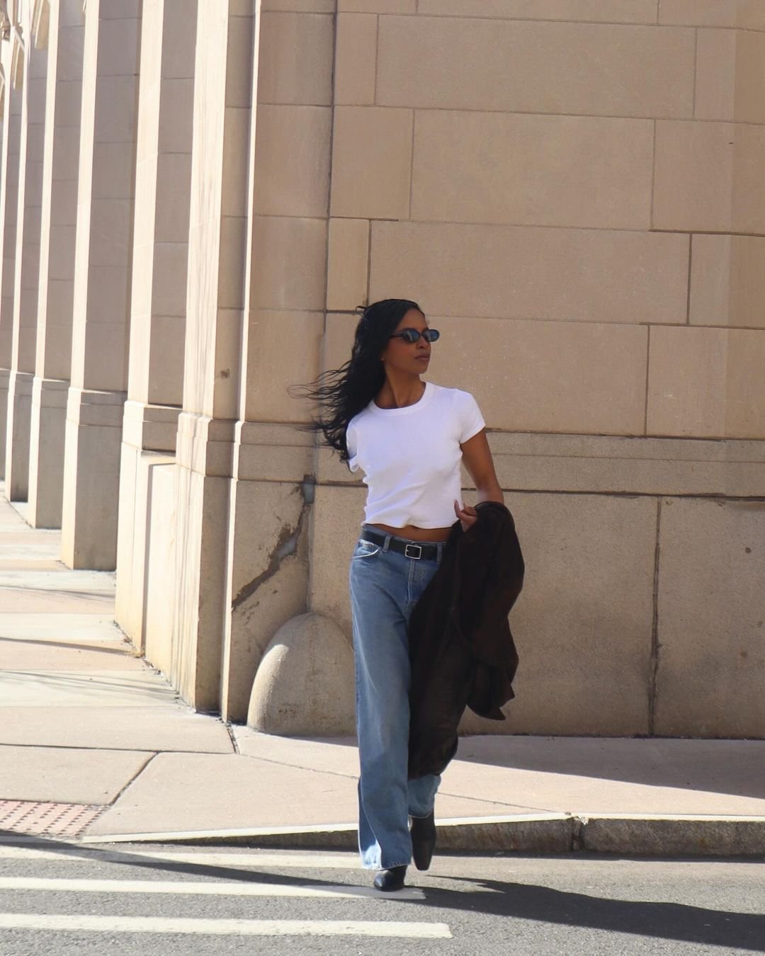 Mulher com camiseta branca básica, jeans de cintura alta e ajustados, cinto preto e botas de bico fino. Ela segura um casaco marrom enquanto caminha, cabelos soltos e óculos de sol. Estilo casual-chic, tendência minimalista.