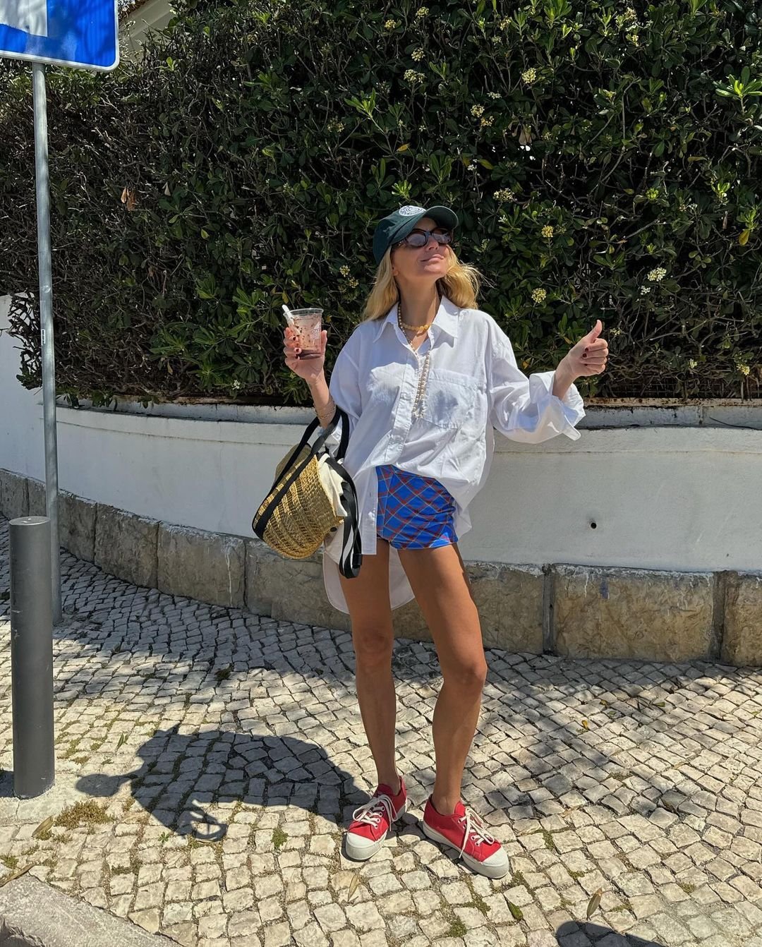 Mulher com estilo casual e despojado, usando camisa branca oversized, shorts estampados, tênis vermelhos e boné verde. Acessórios incluem óculos de sol, colar delicado e bolsa de palha.