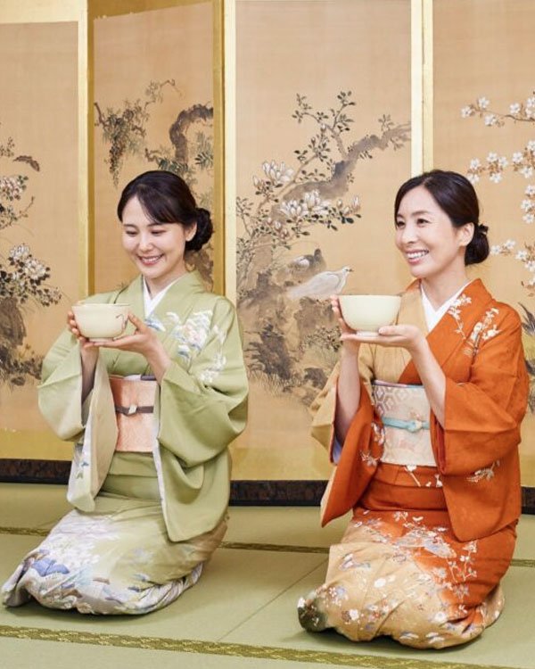 Duas mulheres vestem kimonos tradicionais japoneses, uma em verde claro e outra em laranja, ambas com cintos obi decorados. A cena é de uma cerimônia do chá, refletindo a elegância e a sofisticação da cultura japonesa.