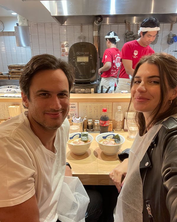 Casal sentado em restaurante de culinária oriental, comendo ramen. Ele veste camiseta branca casual. Ela usa jaqueta de couro preta e blusa branca.