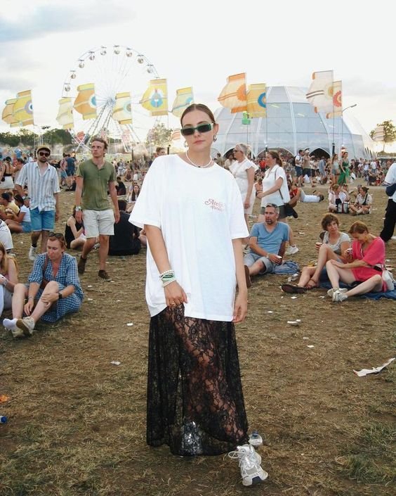 Pessoa em festival ao ar livre, usando camiseta branca oversized, saia preta de renda transparente e tênis esportivos. Acessório inclui óculos de sol pequenos e modernos.