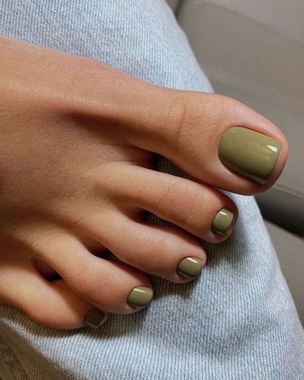 Unhas dos pés pintadas com esmalte verde oliva, tendência minimalista e elegante em pedicures. Dedos dos pés XL. Acompanhadas de calça jeans clara, sugerindo estilo casual e moderno. Ideal para looks versáteis e contemporâneos.