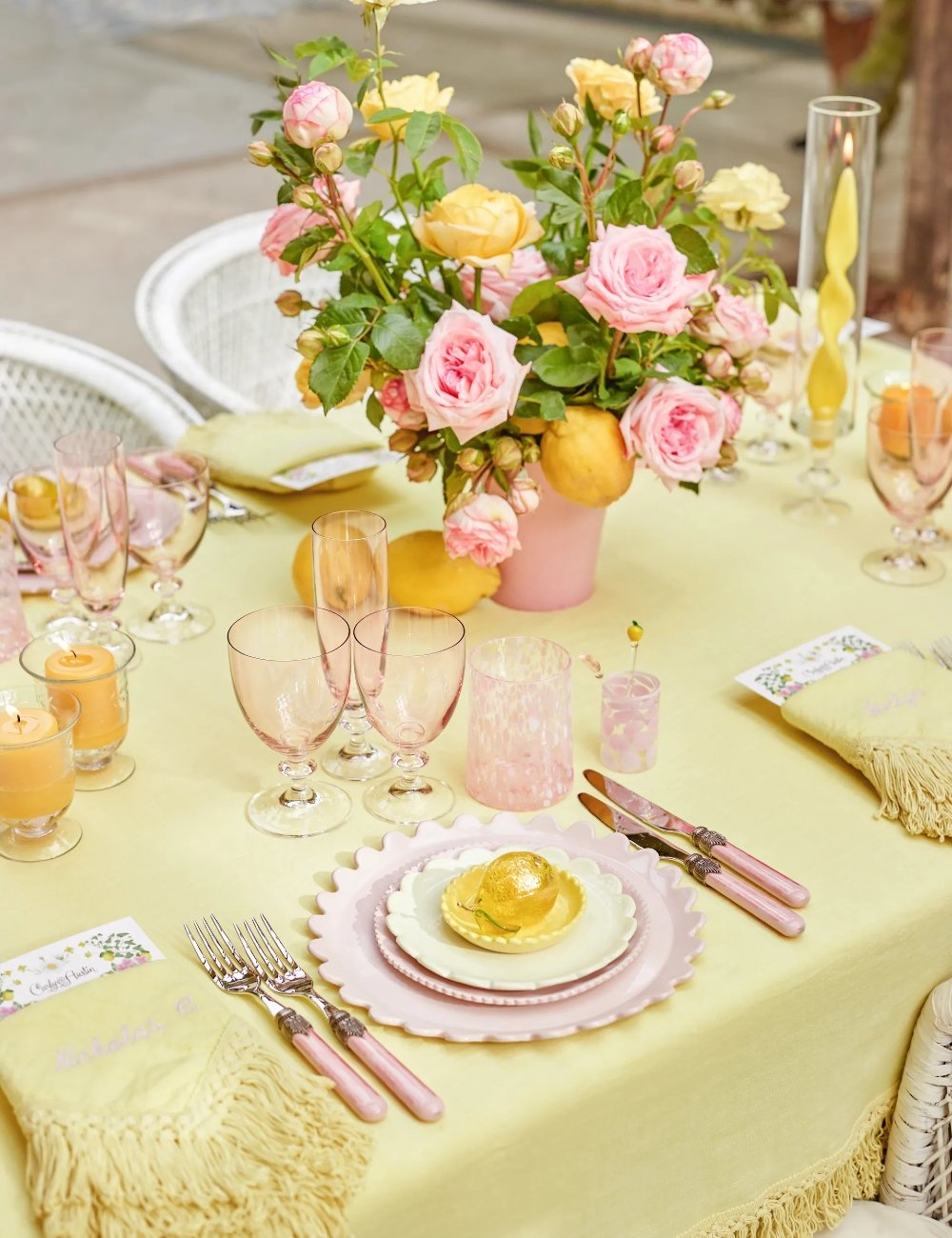 Mesa posta decorada em estilo romântico com toalha de mesa amarela, louça em tons pastel, talheres com cabo rosa, copos delicados e arranjo floral com rosas cor-de-rosa e amarelas. Ideal para ocasiões especiais e encontros elegantes.