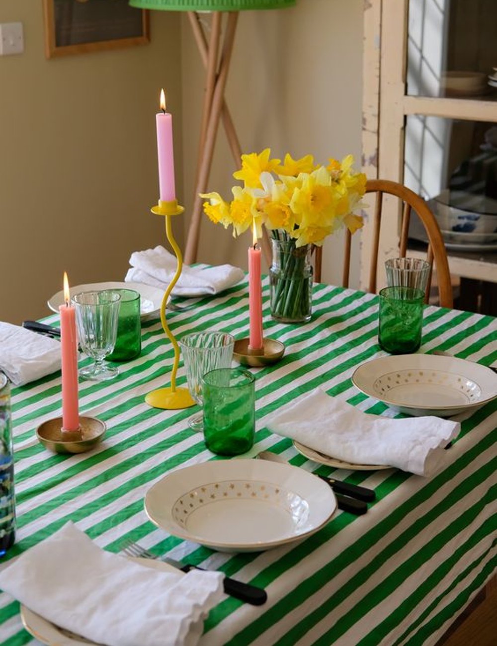 Mesa posta com toalha listrada verde e branca, castiçais coloridos com velas rosa, copos verdes e flores amarelas. Combinação de tendências vintage e color block, com elementos modernos e descontraídos para um ambiente acolhedor e vibrante.