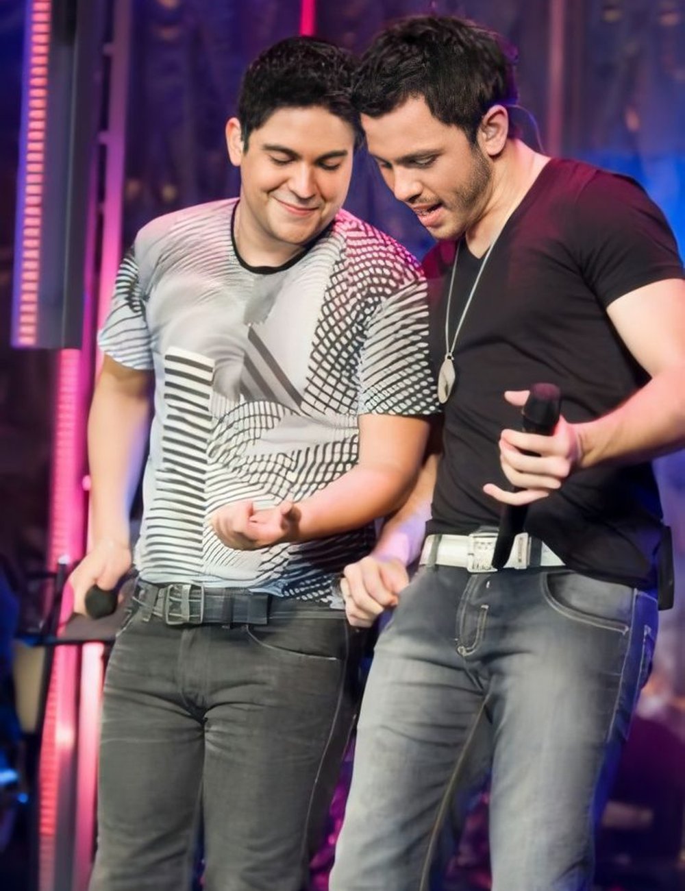 Dois homens em um palco vestem roupas casuais, refletindo tendências modernas. Um usa camiseta estampada com padrões geométricos e jeans escuros, enquanto o outro veste camiseta preta básica e jeans desbotados. Sertanejo.