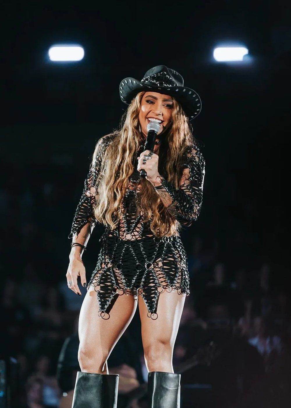 Cantora veste look ousado com um vestido de rede de malha preta, chapéu estilo cowboy e botas de cano alto. O visual mistura tendências country e modernas, destacando-se pela combinação de elementos rústicos e fashion.