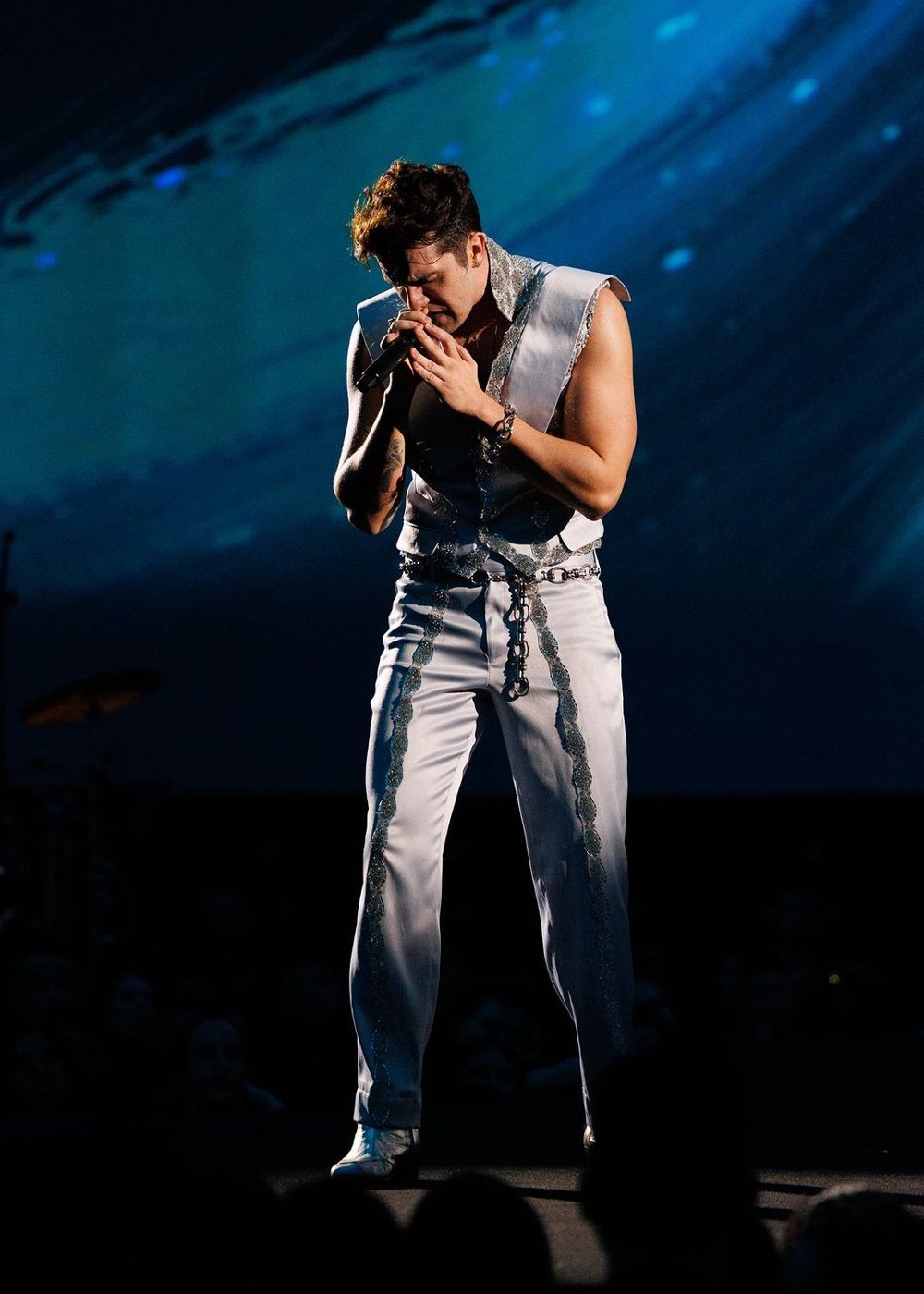 Cantor com estilo ousado no palco, veste colete prata sem mangas e calça combinando com detalhes brilhantes. O look é complementado por correntes e sapatos metálicos, refletindo tendências futuristas e glam-rock na moda masculina.