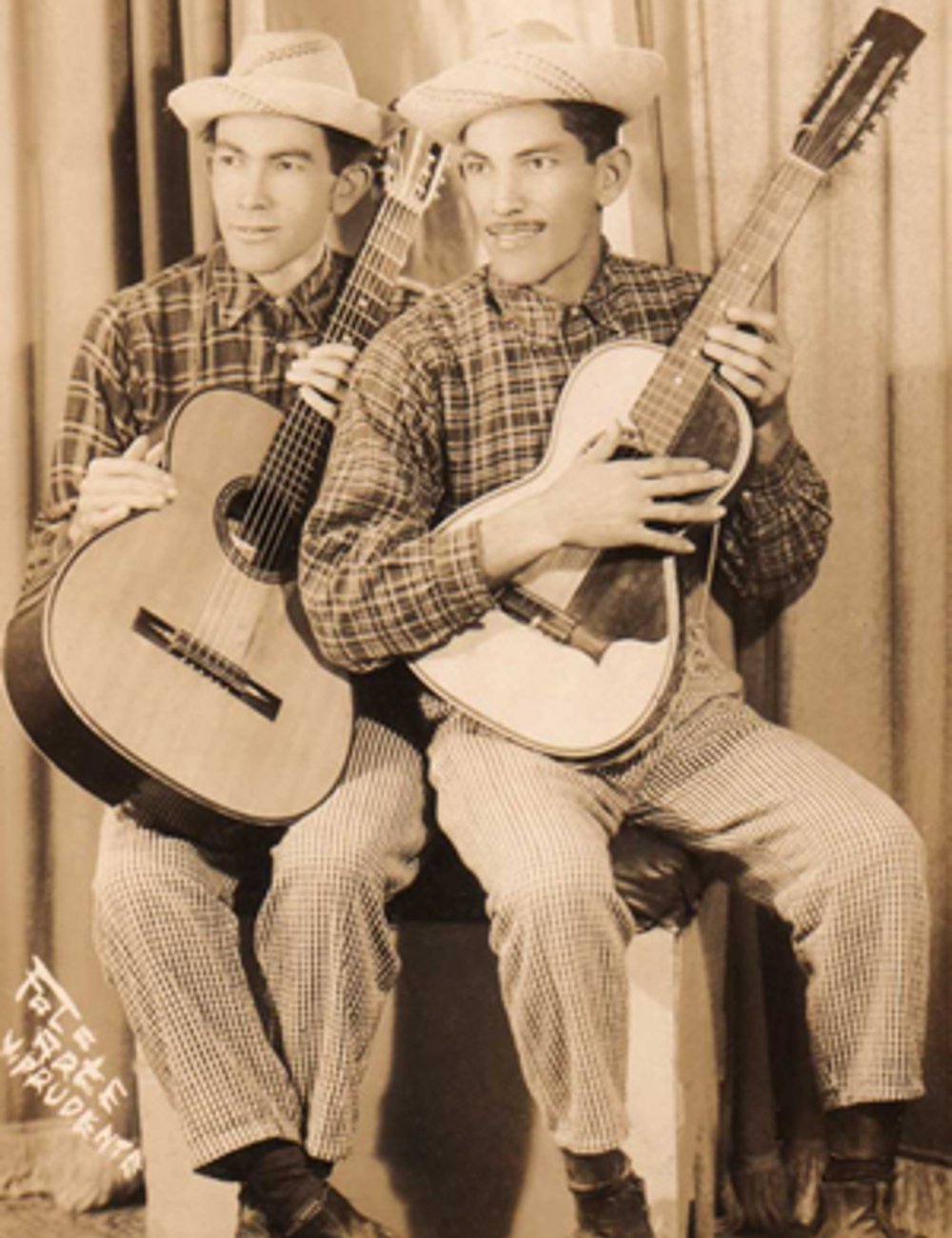 Homens vestindo camisas xadrez de mangas longas e calças quadriculadas posam com violões. Eles usam chapéus de palha, representando um estilo típico de sertanejo. Cortinas ao fundo indicam um ambiente de estúdio.