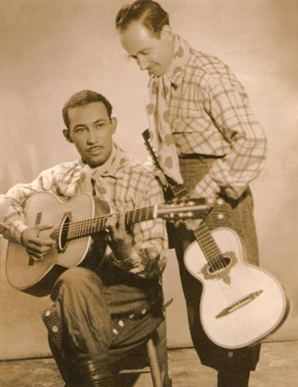 Imagem em preto e branco de dois homens usando roupas tradicionais de gaúchos. Ambos estão vestindo camisas xadrez com lenços no pescoço, calças bombachas e botas. Um deles está tocando violão, sertanejo.