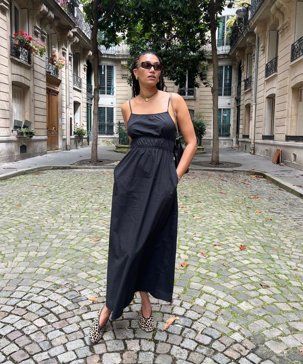 Mulher elegante vestindo um vestido preto longo e solto com alças finas em um cenário urbano clássico. Ela combina o look com sapatos de estampa animal, óculos de sol e acessórios dourados minimalistas, destacando um estilo moderno e sofisticado.