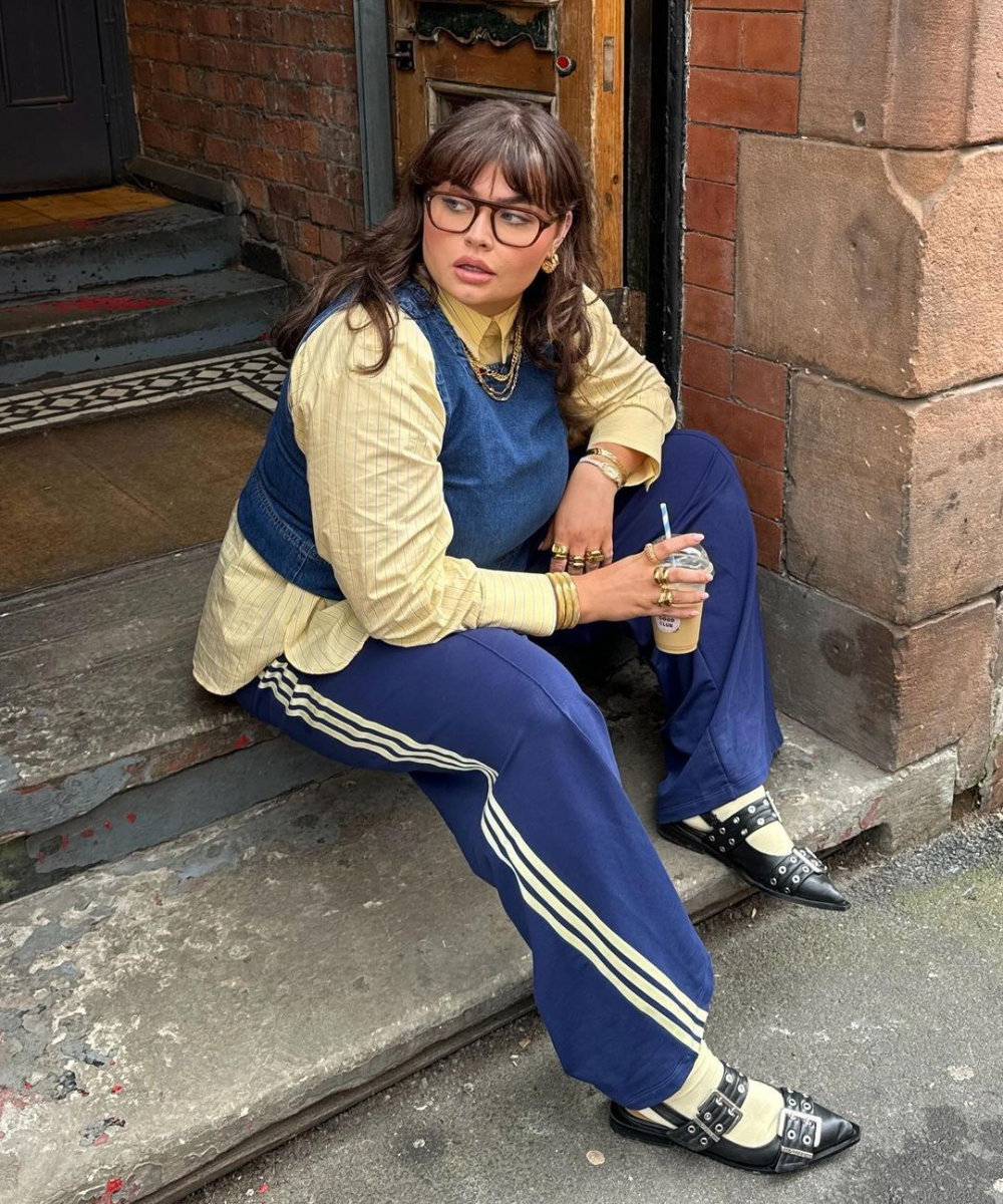 Mulher sentada na escada usa camisa amarela listrada, colete jeans, calça azul esportiva com listras brancas e sapatos de fivela pretos com meias claras. Ela usa óculos, brincos, colares e vários anéis dourados, segurando uma bebida. Estilo combina v