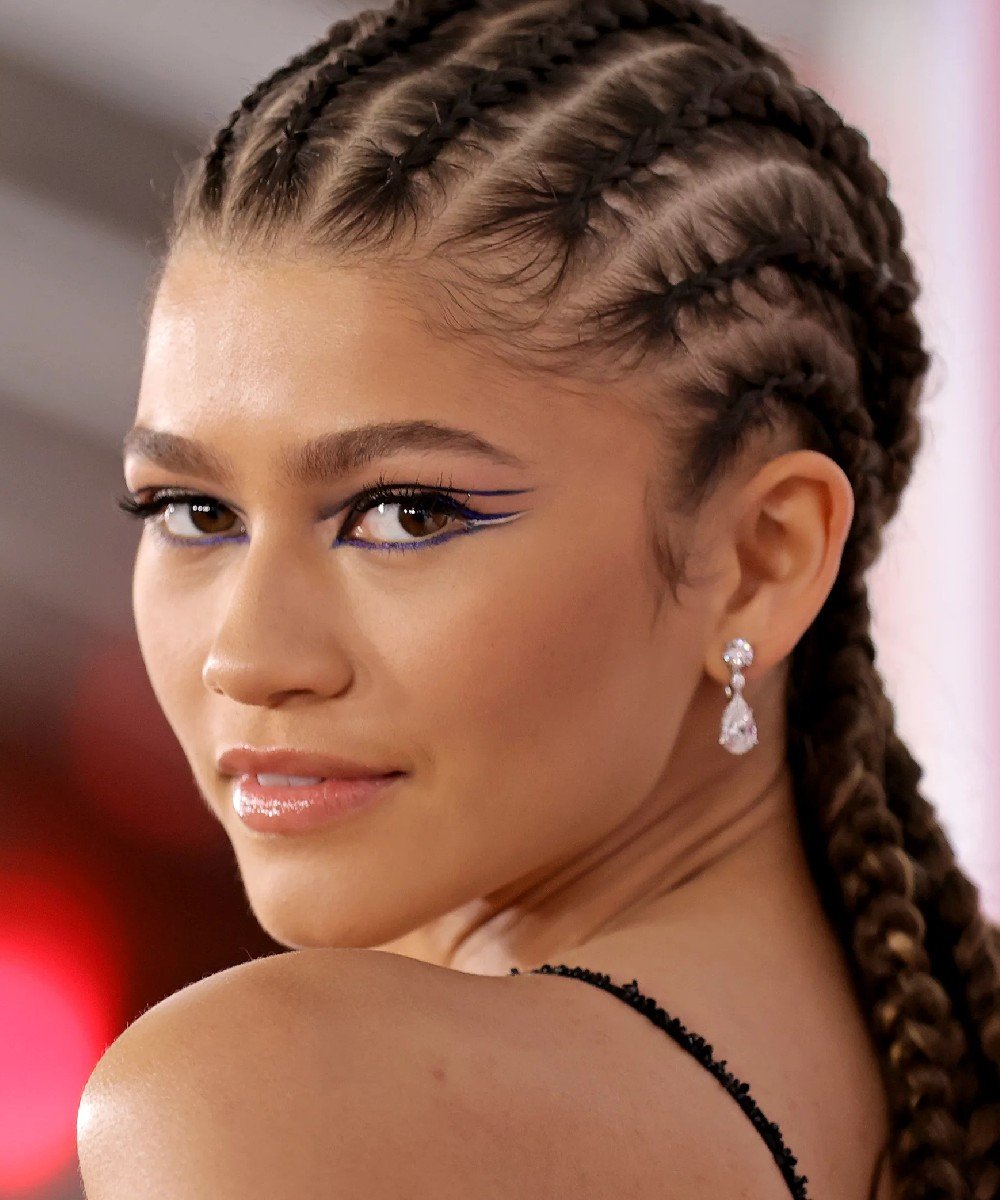 Zendaya usando tipo de trança nagô, maquiagem ousada com delineado gráfico azul, brincos pendentes e estilo elegante. Tranças nagô são tendência em penteados protetores, enquanto a maquiagem destaca a criatividade nos olhos.