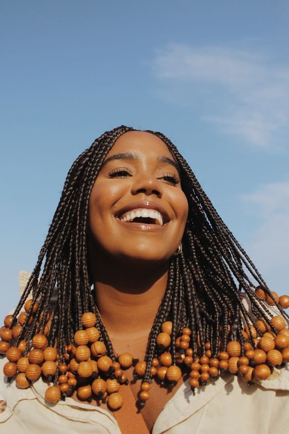 Mulher sorridente usando box braids com missangas ao ar livre. A roupa neutra complementa o visual ousado e moderno das tranças.