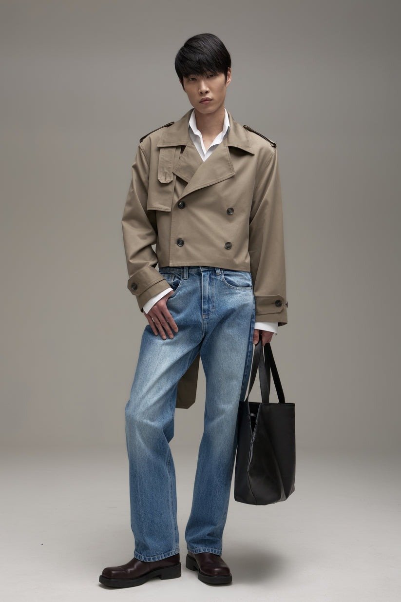 Modelo vestindo trench coat bege, calça jeans de lavagem clara e sapatos marrons. Estilo casual e sofisticado, combinando elementos clássicos e contemporâneos. A bolsa preta adiciona um toque funcional, refletindo tendências de moda urbana atual.