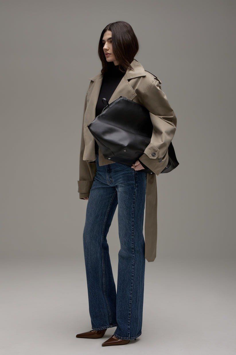 Mulher vestindo trench coat bege com blusa preta de gola alta, combinando com jeans de corte reto e sapatos de bico fino marrom. Carrega uma bolsa grande preta de couro. Look contemporâneo que mistura elegância e casualidade, ideal para o outono/inverno.