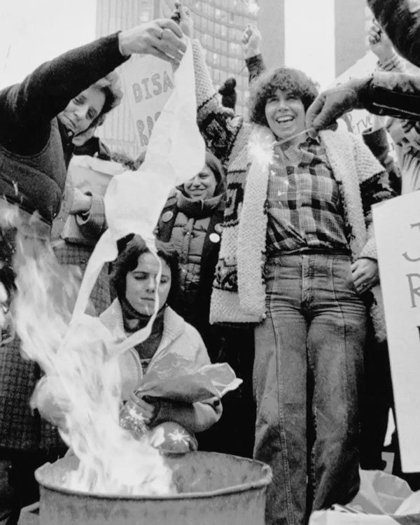 Manifestantes com vestuário típico dos anos 70, incluindo camisas de flanela xadrez e jaquetas de lã grossas. Moda casual e confortáveis, refletindo um estilo despojado e rebelde da época, evidenciando a expressão de liberdade pessoal e social.