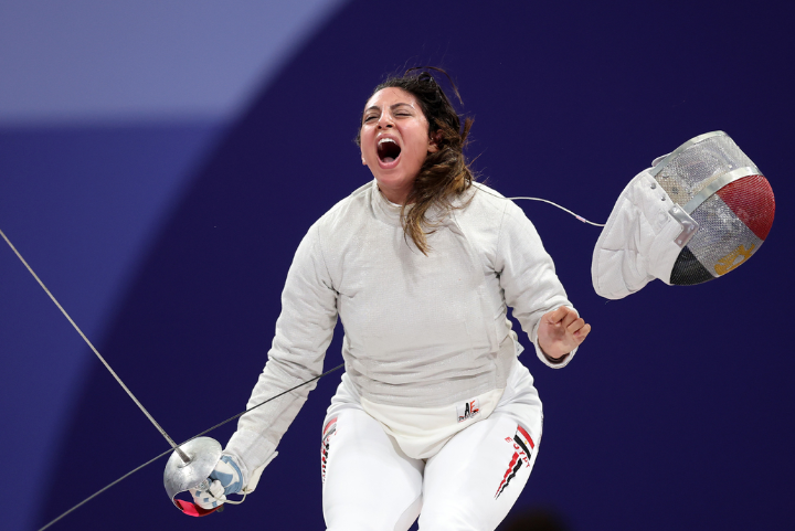 Imagem de uma esgrimista em ação, vestindo uniforme completo de esgrima: jaqueta branca acolchoada, calça branca justa com detalhes laterais e máscara de proteção removida. O look destaca a combinação de proteção e performance esportiva.