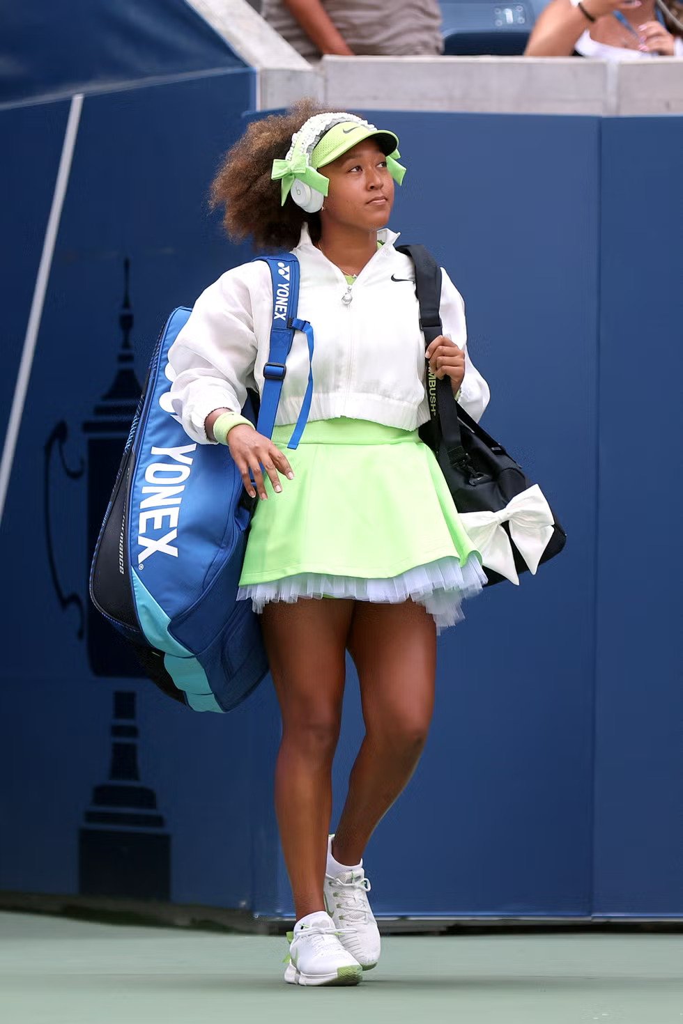  Naomi Osaka Tenista vestindo um conjunto esportivo estiloso e moderno, com saia verde neon, jaqueta branca cropped, tênis brancos e viseira verde combinando. Acessórios incluem fones de ouvido brancos e uma grande bolsa Azul Yonex. Estilo que combina conforto e performance.