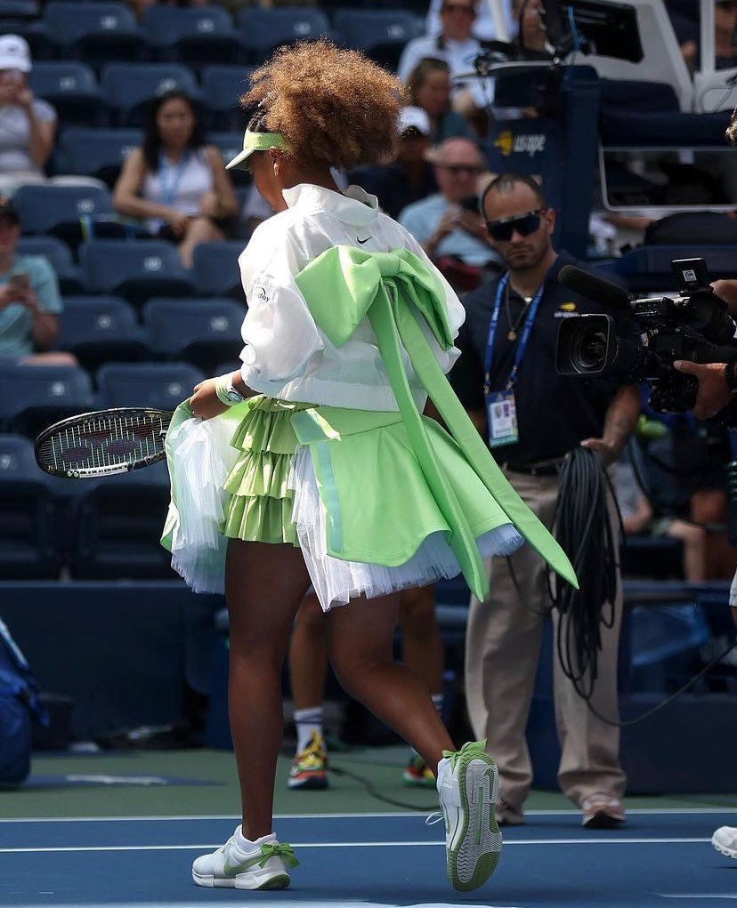  Naomi Osaka Jogadora de tênis veste conjunto moderno em tons de verde e branco, com saia plissada, jaqueta leve e tênis esportivos. O look destaca-se pela pegada futurista e uso de cores vibrantes, combinando funcionalidade esportiva com estilo. tendências de moda contemporânea.