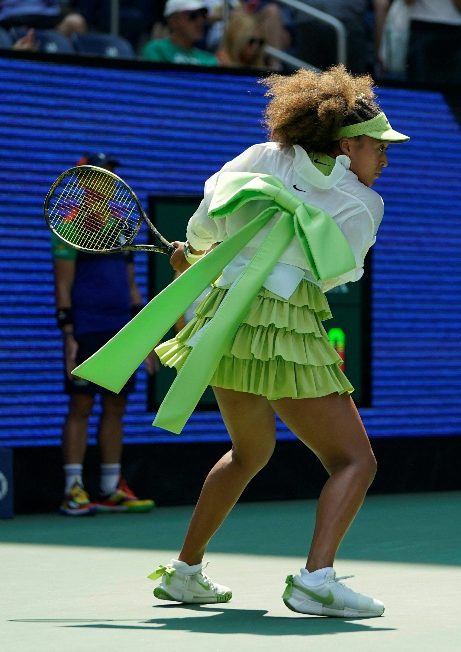 Naomi Osaka Jogadora de tênis vestindo saia plissada verde-clara e blusa branca com laço verde chamativo nas costas. Complementa o look com tênis brancos decorados com laços verdes e boné esportivo verde. Estilo esportivo e elegante, marcando tendência em partidas de tênis.